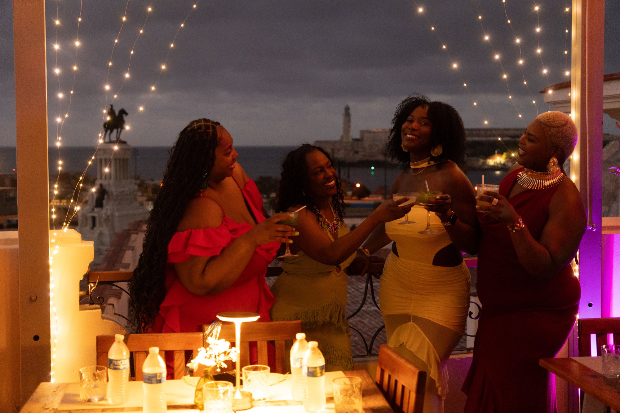 Somewhere with a view and music — four friends toasting with cocktails on a rooftop at dusk, string lights above, El Morro lighthouse across the bay behind them