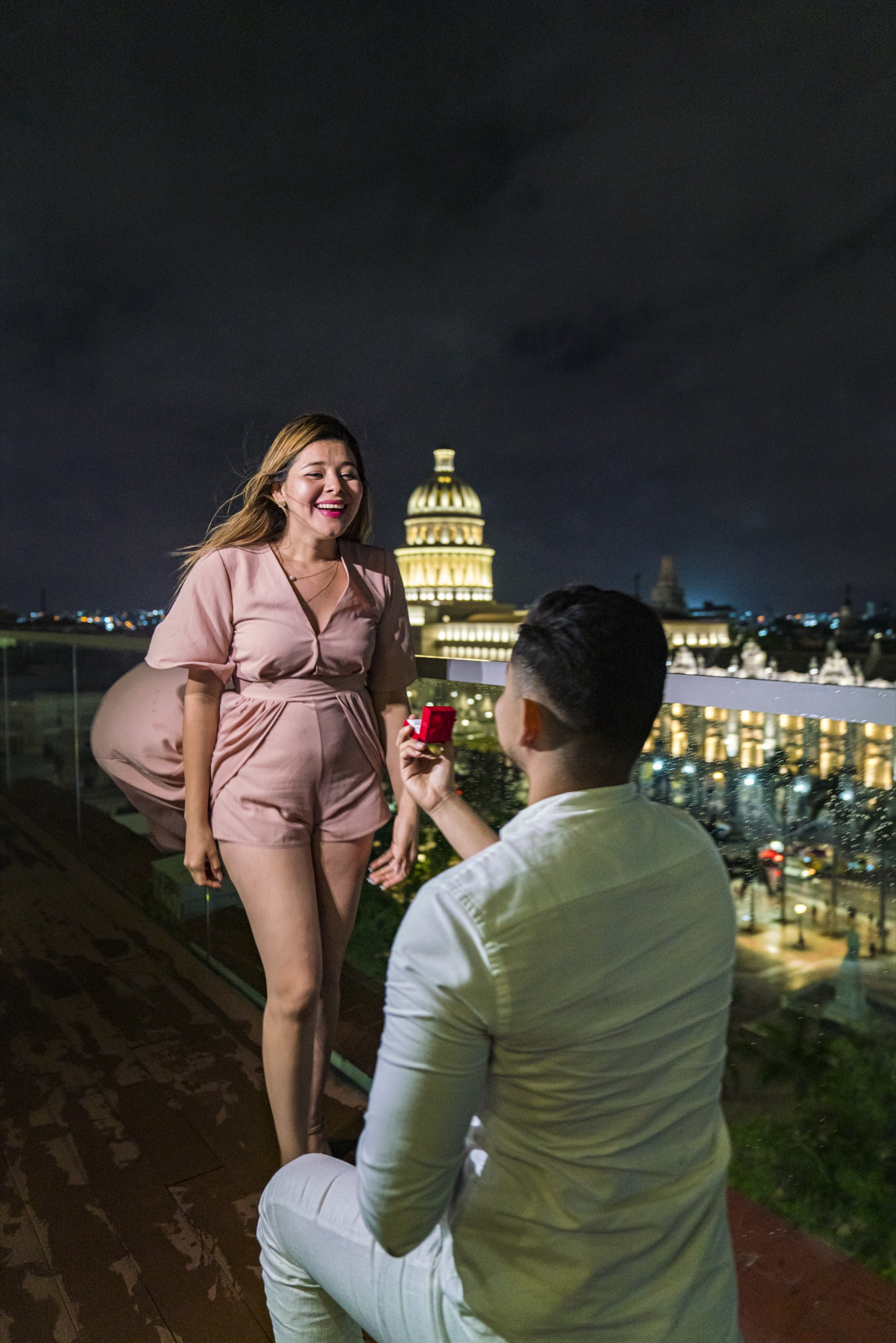 A man proposing on a Havana rooftop at night, his partner laughing with joy, the illuminated Capitolio dome glowing behind them