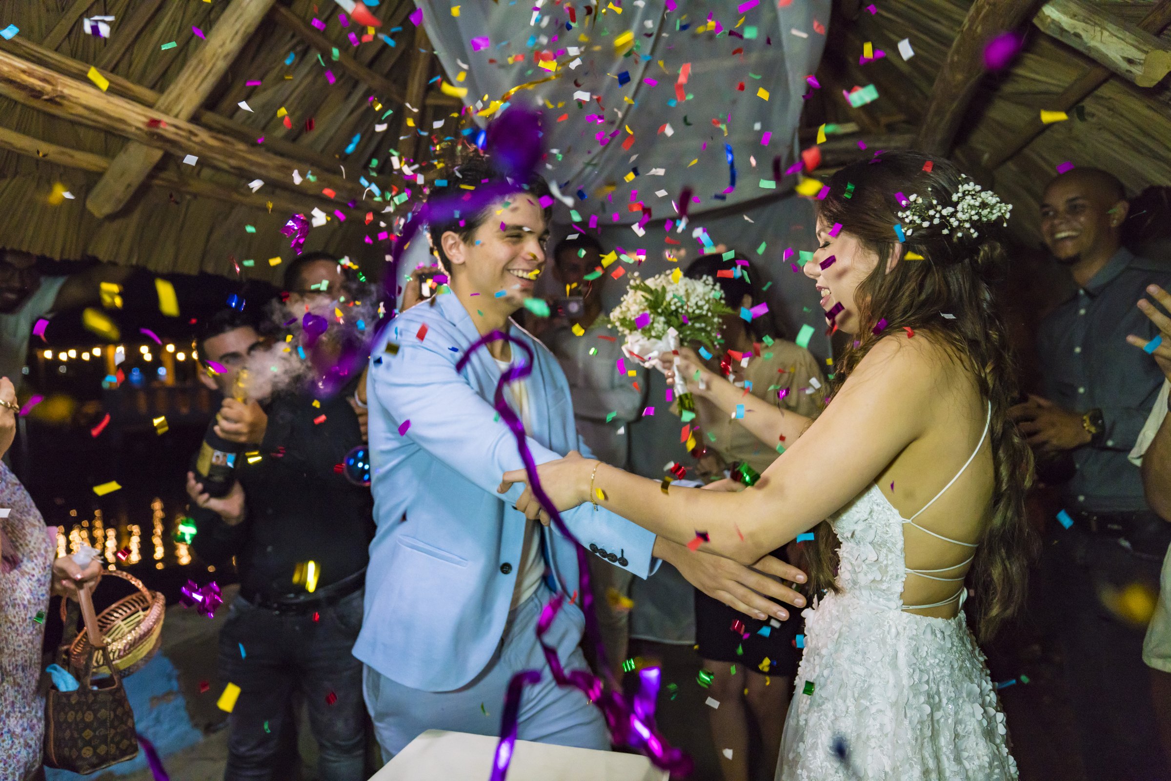 A bride and groom laughing under a shower of confetti at their reception, guests cheering around them under a thatched roof