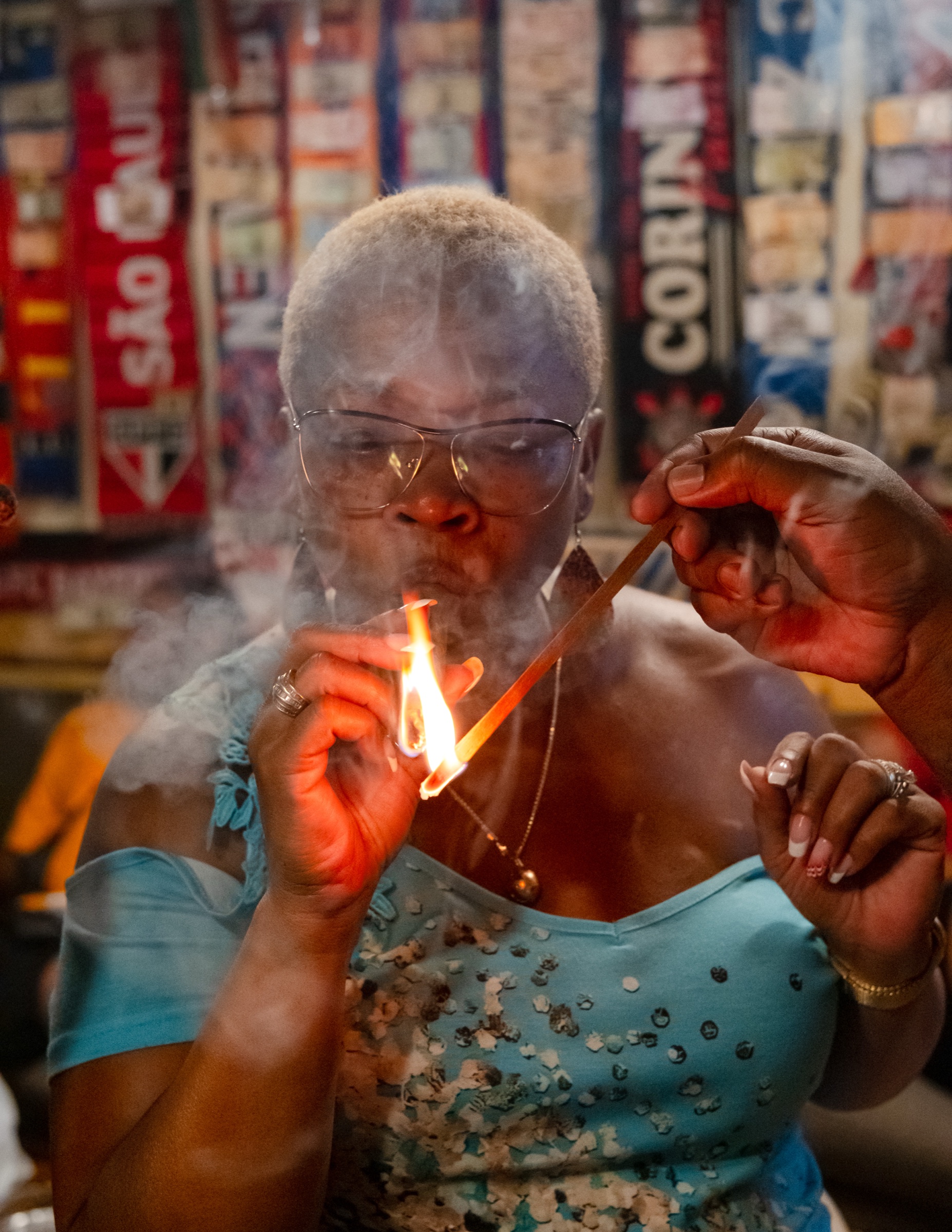 A woman in a turquoise dress carefully lighting a cigar, the flame and a curl of smoke lit warm against the dark interior of a Havana shop