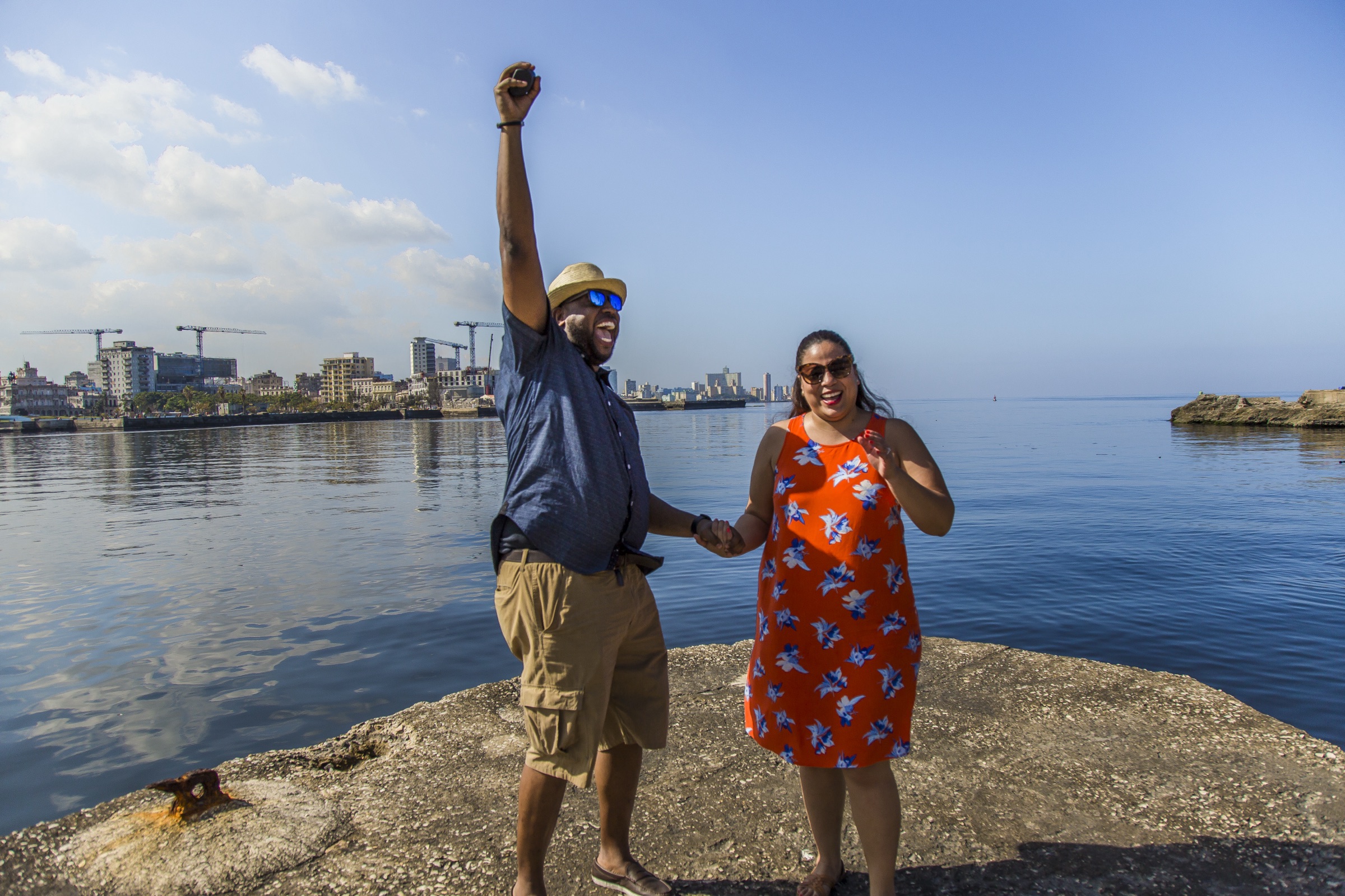 A newly engaged couple celebrating on the rocks by the water, the man's fist raised in the air, the Havana skyline behind them