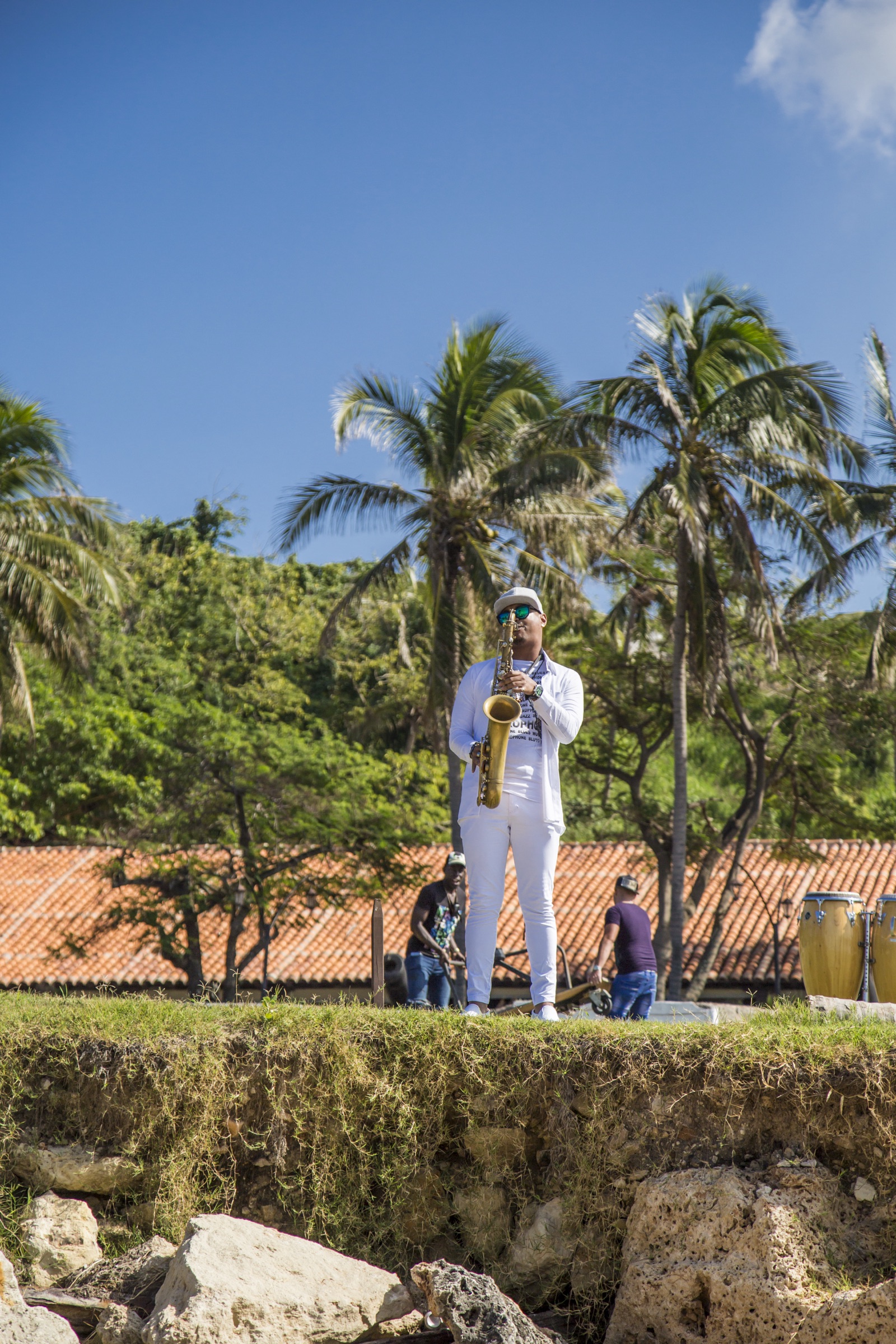 A Cuban musician in white playing saxophone on the grass, palm trees overhead and the band's congas set up behind him