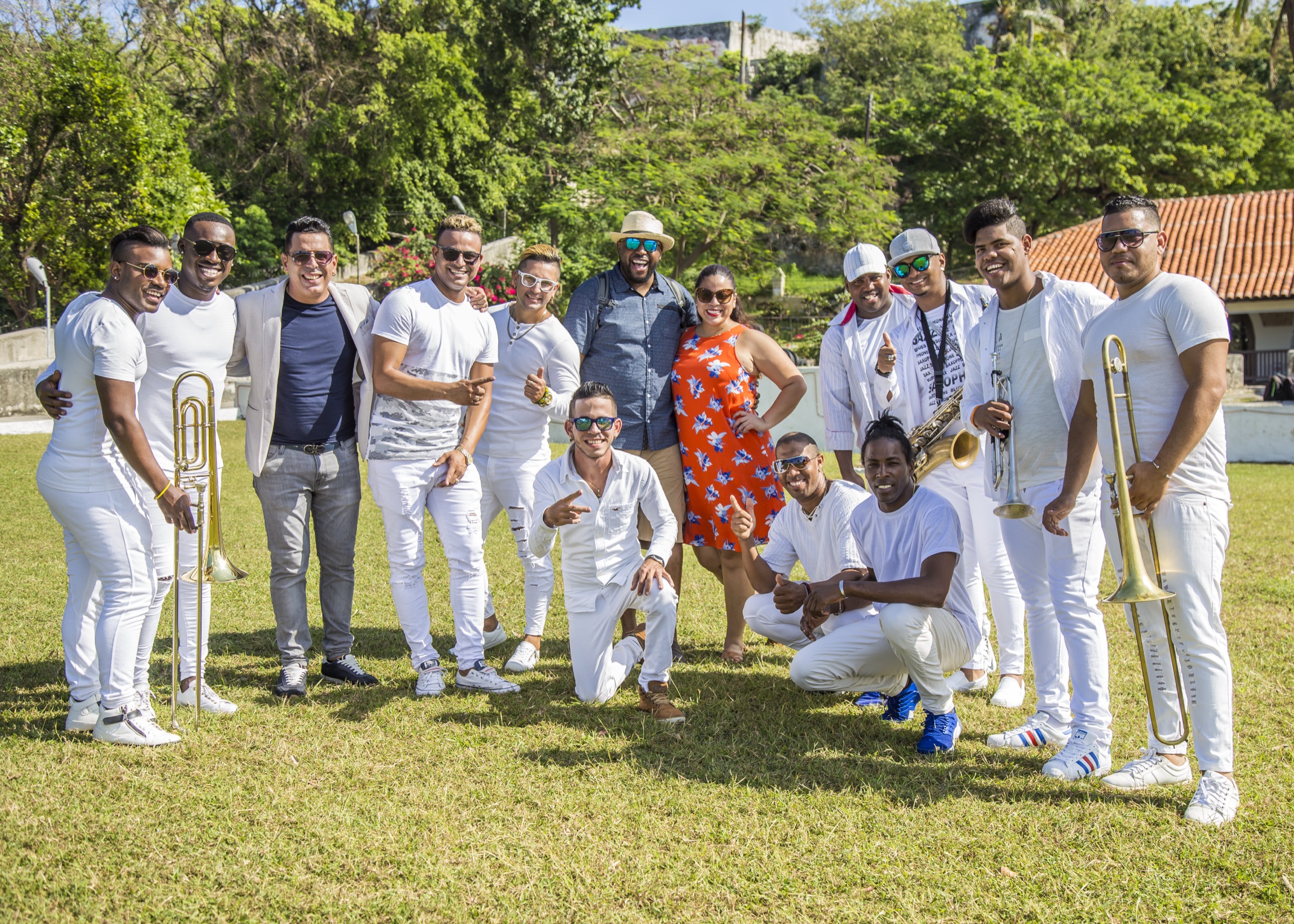 A full Cuban band dressed in white, with trombones and a saxophone, posing on the grass with a newly engaged couple