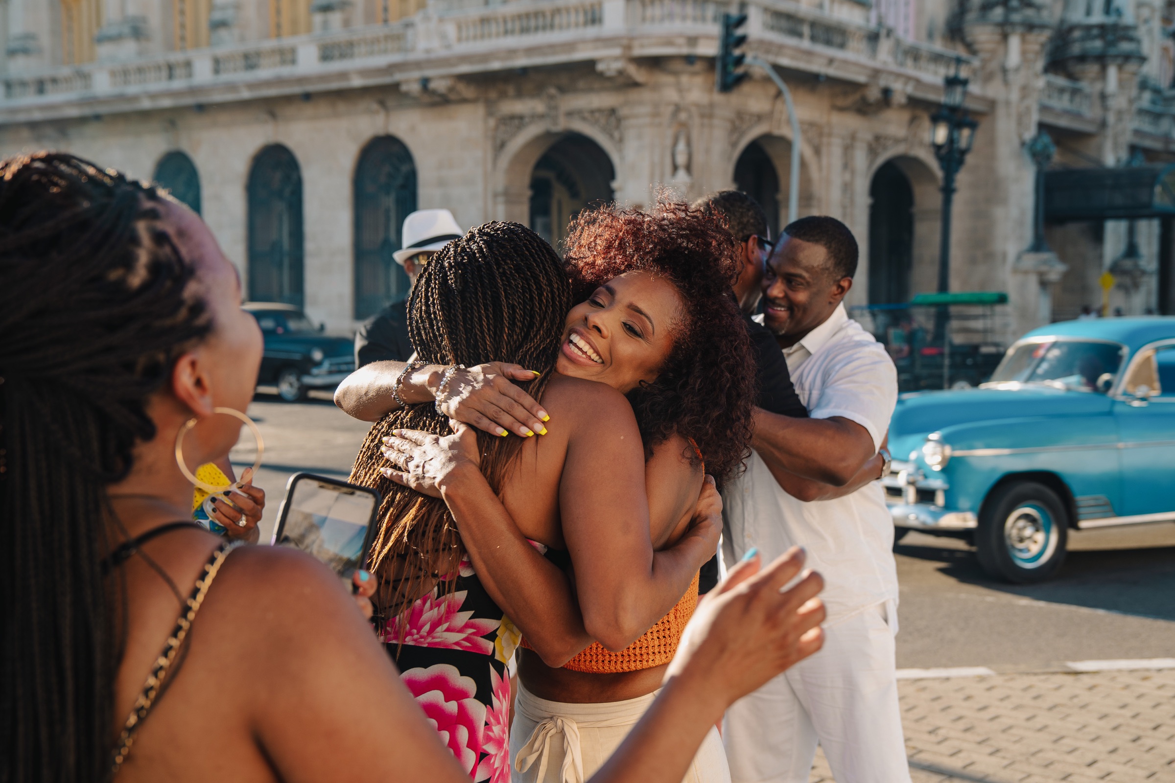 Friends rushing in to hug a newly engaged couple on a Havana street, a classic car passing behind them