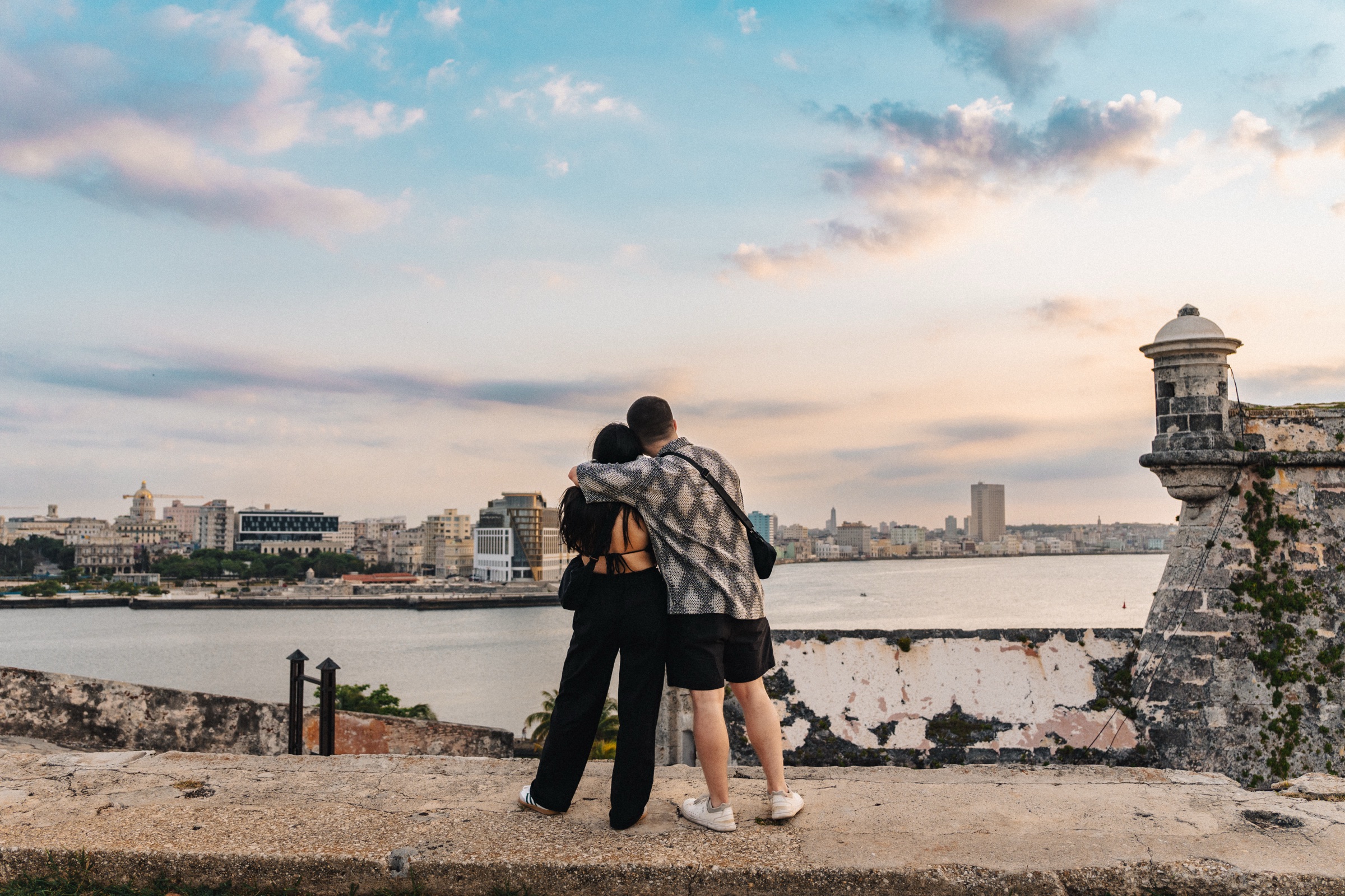 A couple embracing at a fortress overlook at dusk, looking out at the Havana skyline across the bay