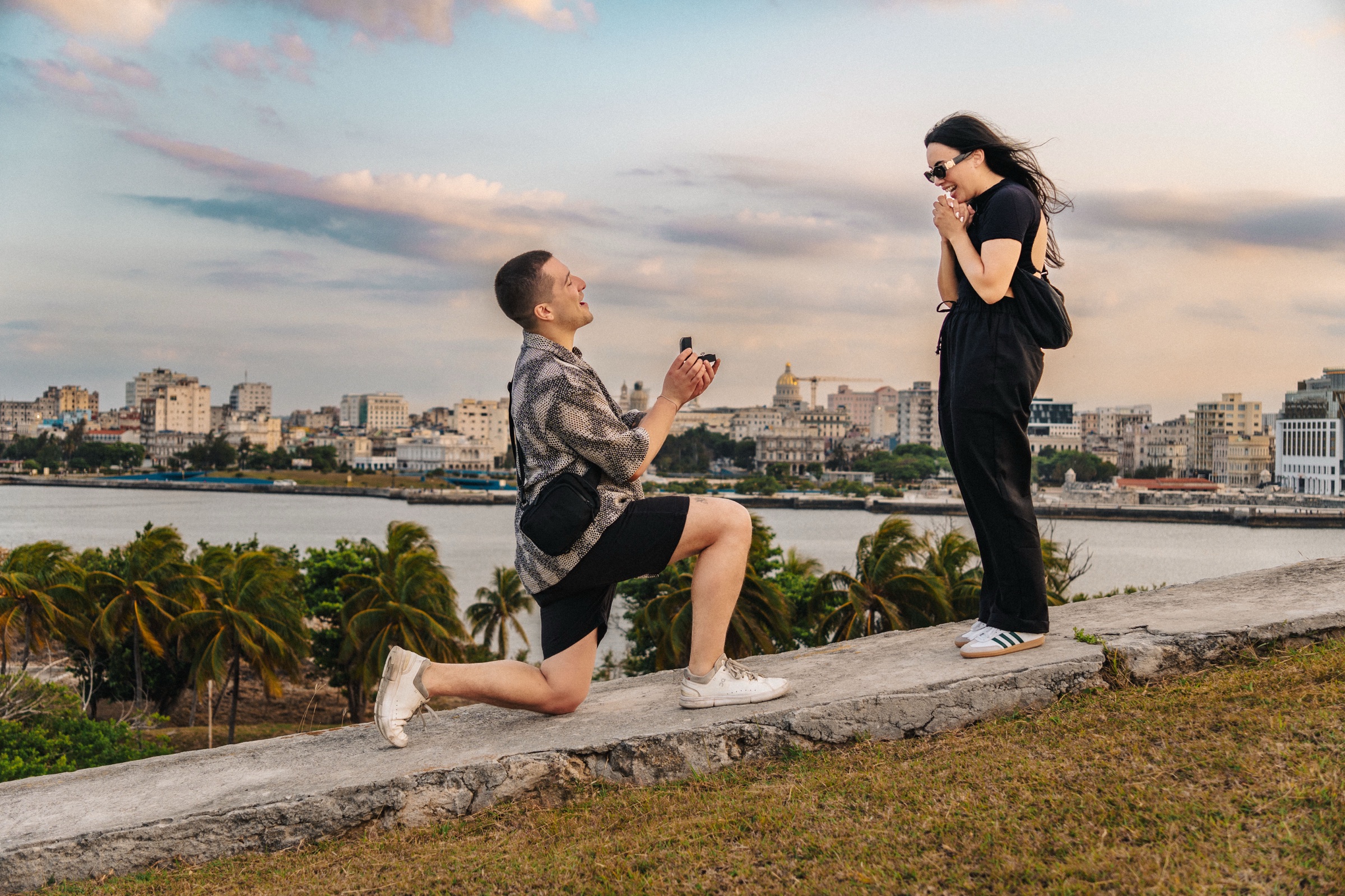 A man kneeling on a low wall holding out a ring, the woman covering her face in shock, with palm trees and the Havana skyline behind them at dusk