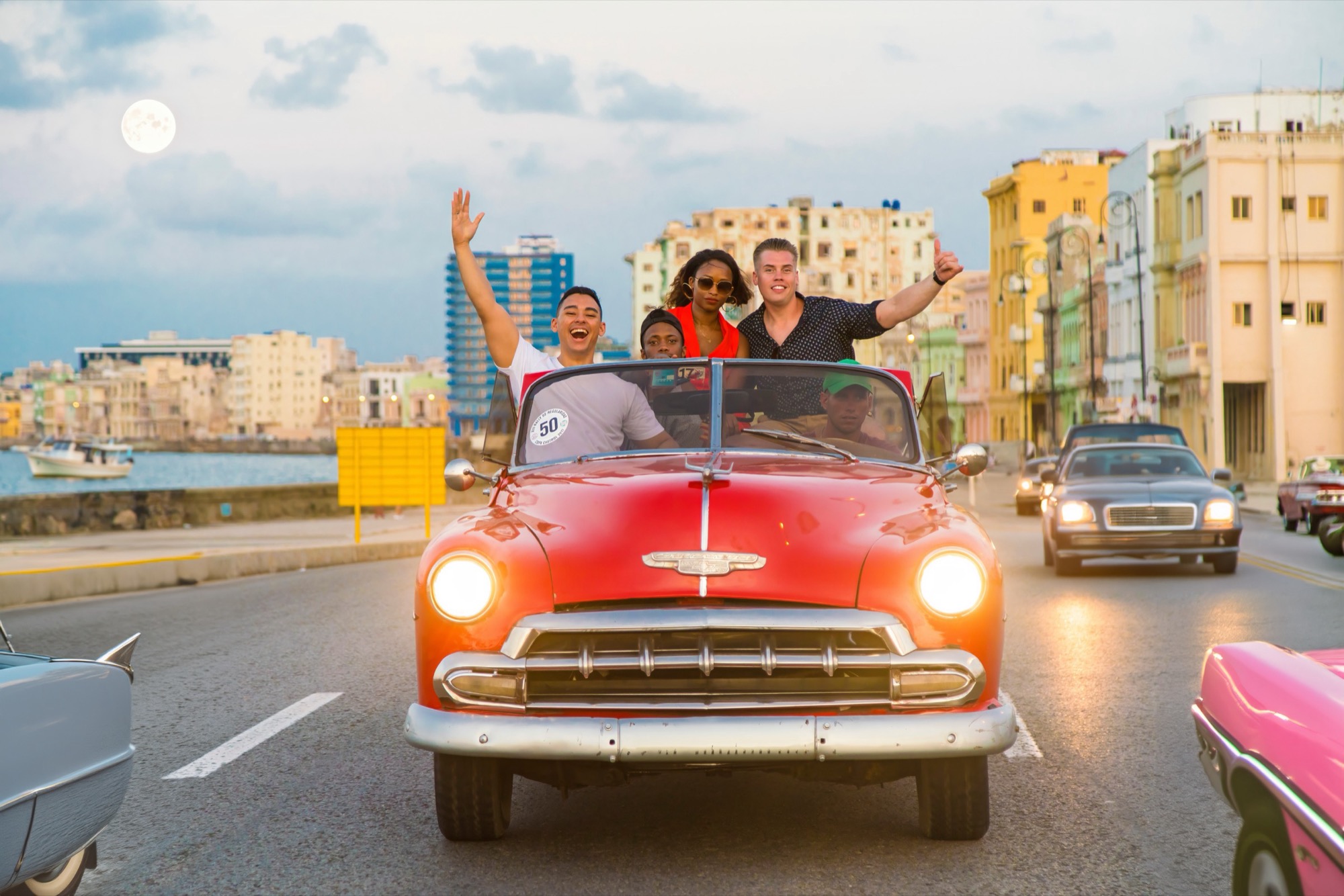 A group riding a red classic convertible down the Malecón, arms raised