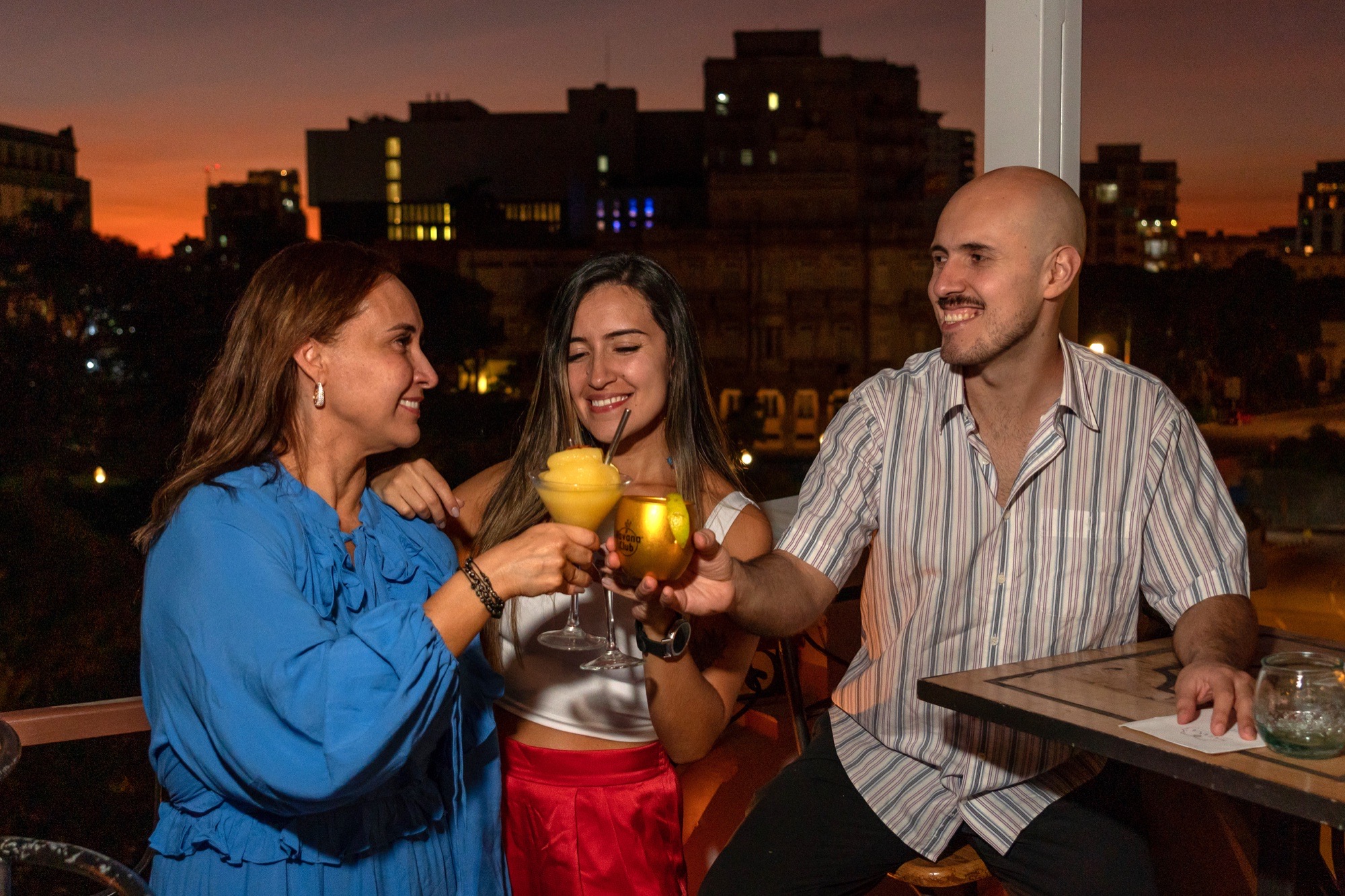 Three friends toasting cocktails on a rooftop as the sun sets over Havana