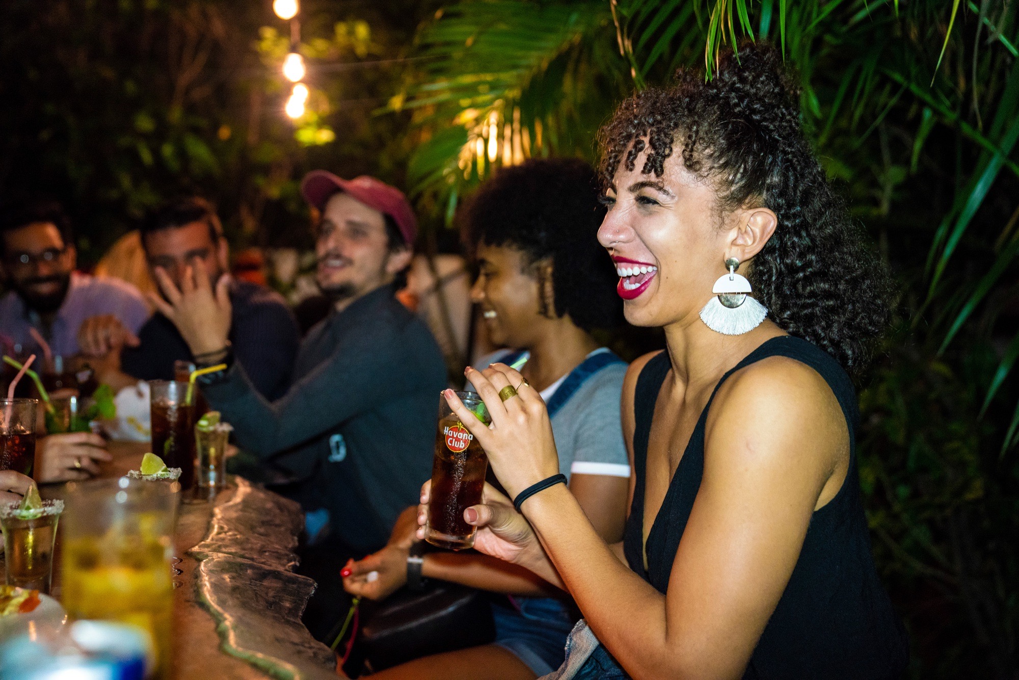 A laughing crowd with cocktails at a warmly lit Havana bar at night