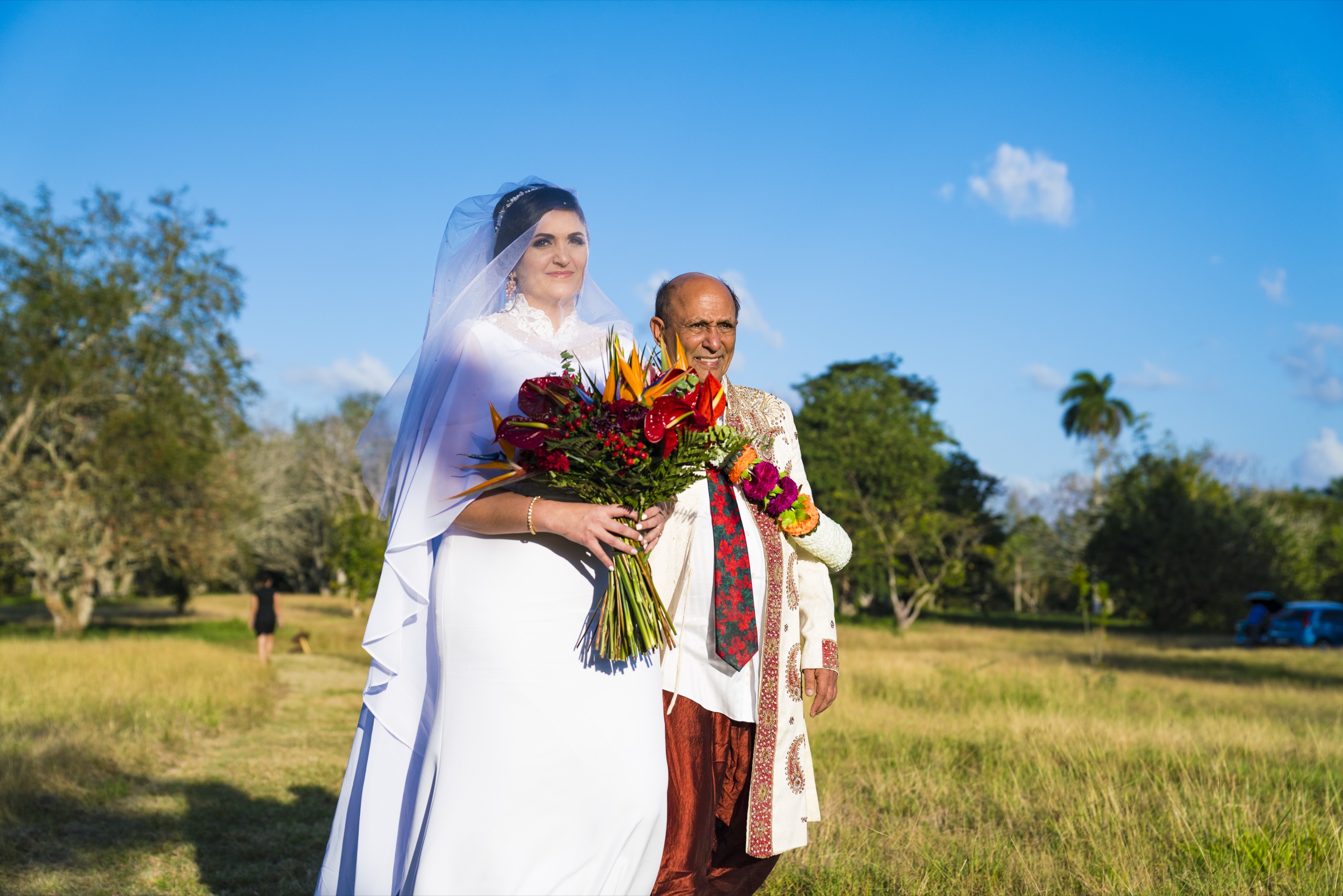 The bride in a white gown and veil holding a bouquet of tropical flowers, walking through a golden field beside her father in an embroidered sherwani