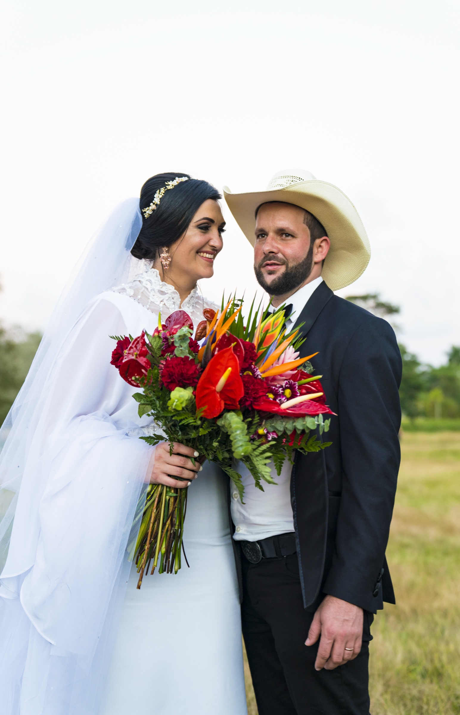 The bride in white lace and veil smiling beside the groom in a black suit and pale cowboy hat, holding a bouquet of red and orange tropical flowers in an open field
