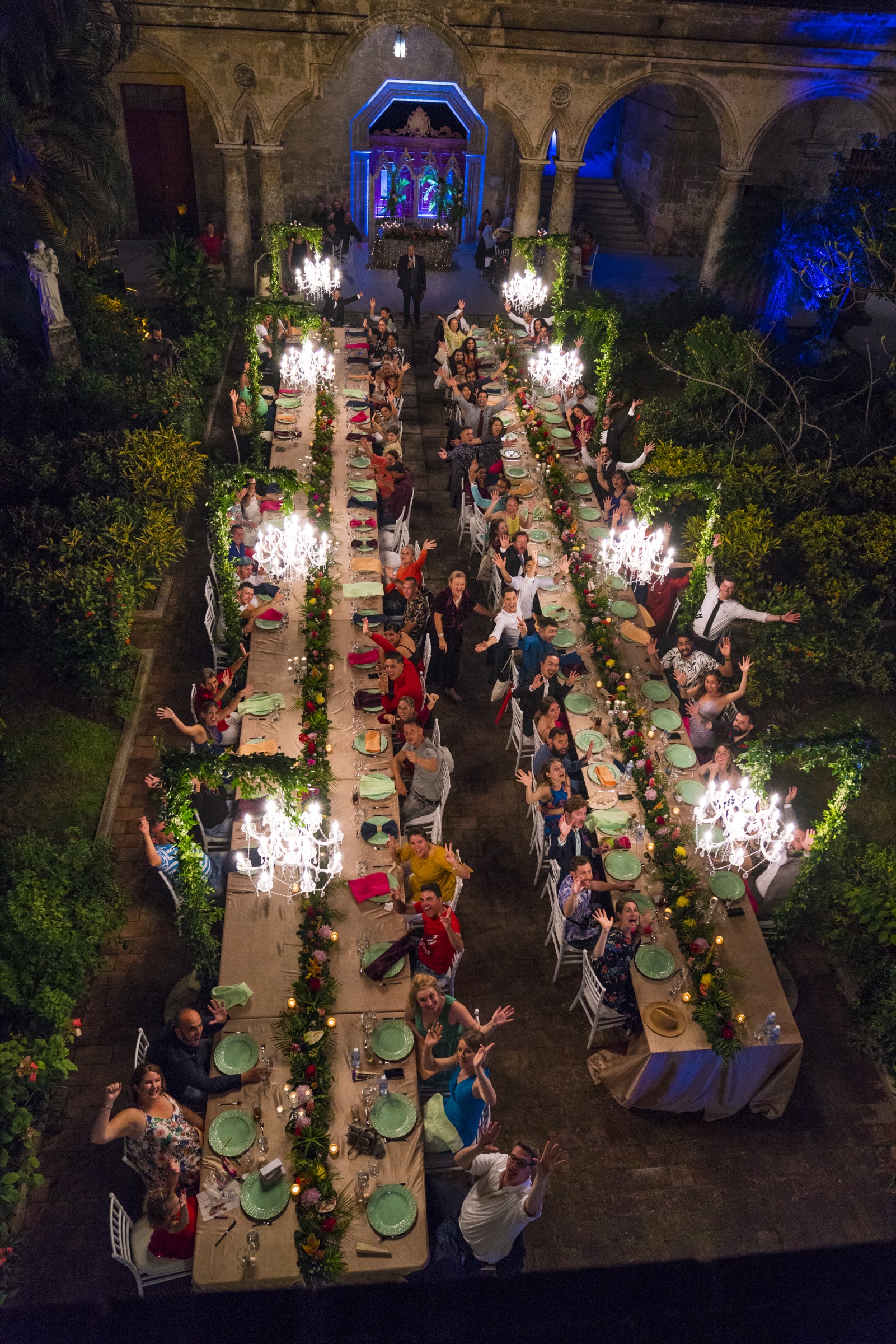 A nighttime aerial view of two long banquet tables full of guests in a colonial courtyard, lit by chandeliers and lined with greenery and flowers