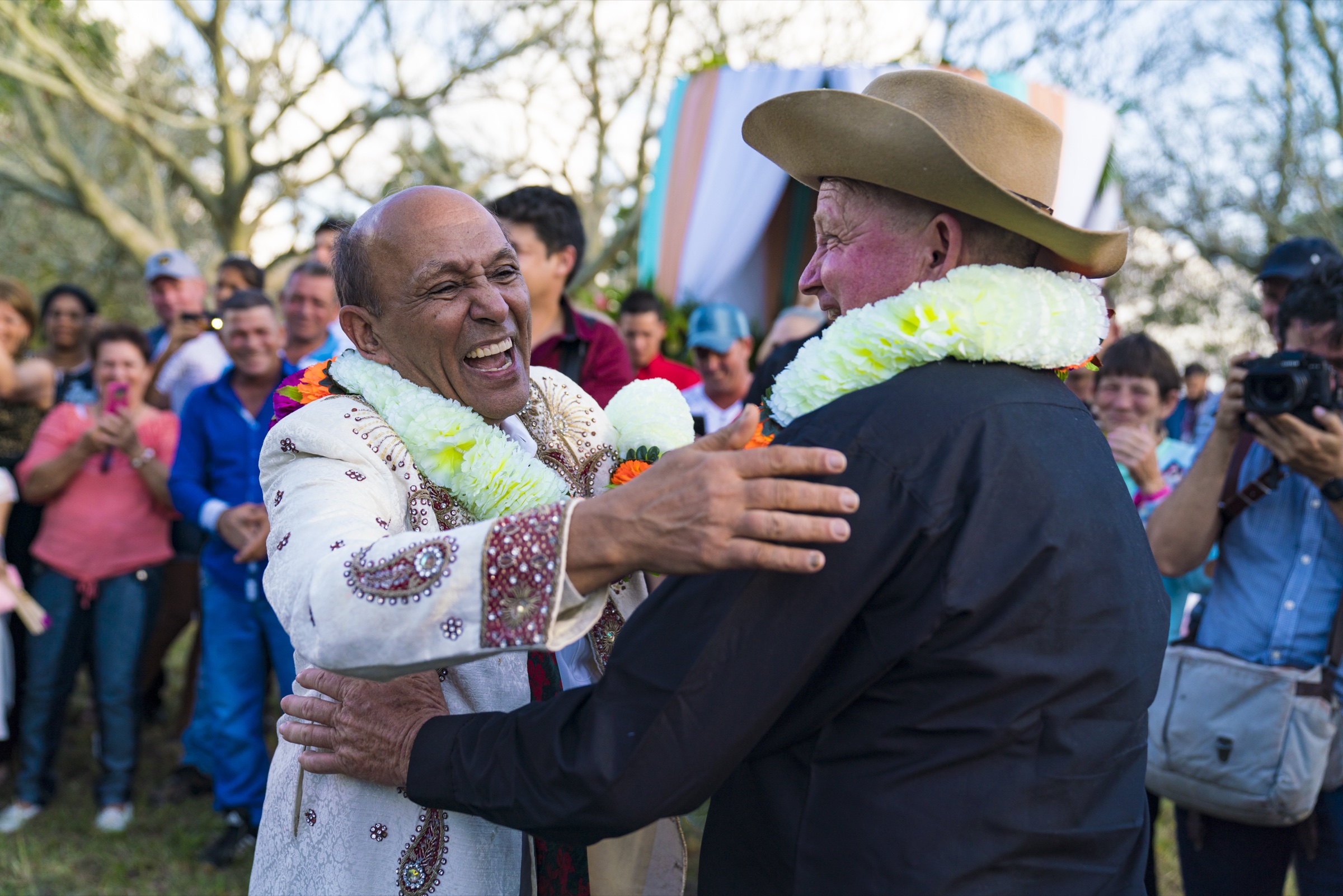 The bride's father in an embroidered sherwani and the groom's father in a wide country hat embracing and laughing, both wearing flower garlands, with the wedding guests applauding behind them