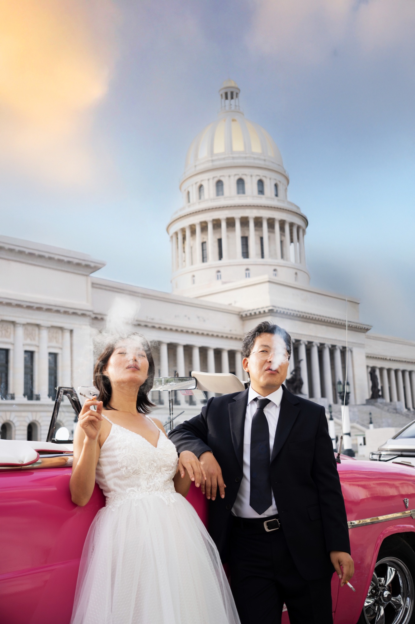 A bride and groom leaning on a pink classic convertible in front of the Capitolio