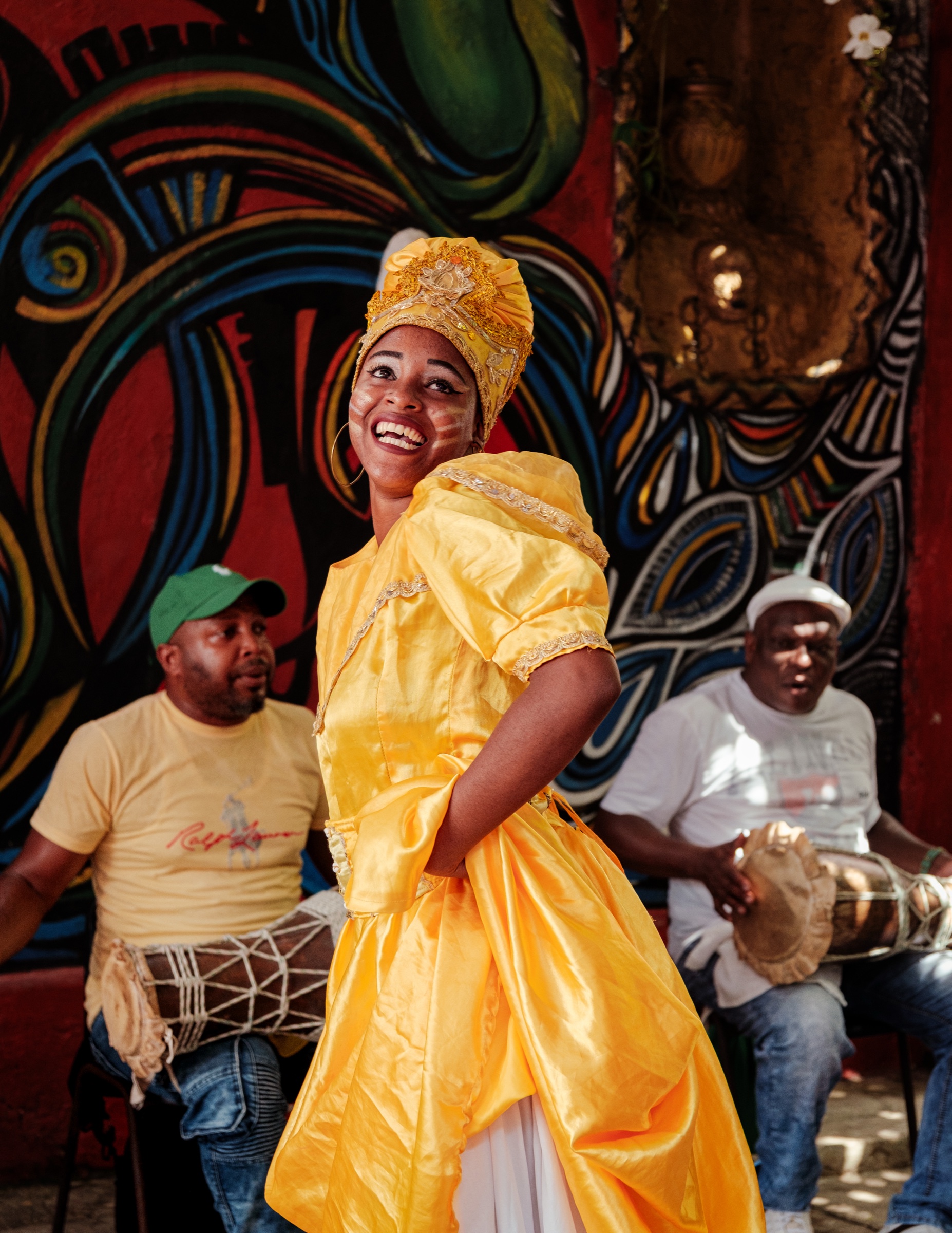 An Afro-Cuban rumba dancer in a bright yellow dress laughing, drummers playing behind her against the painted Hamel murals