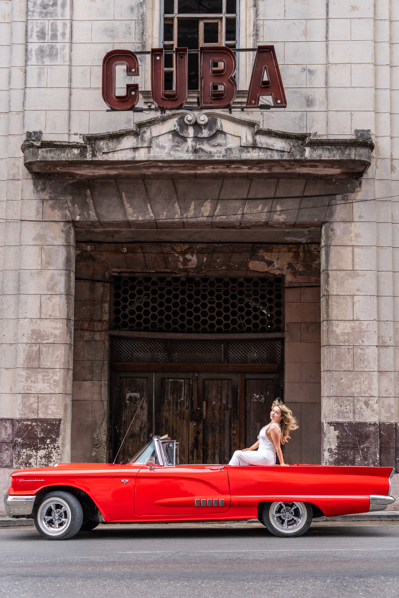 A woman in white on a red convertible below the giant CUBA cinema sign
