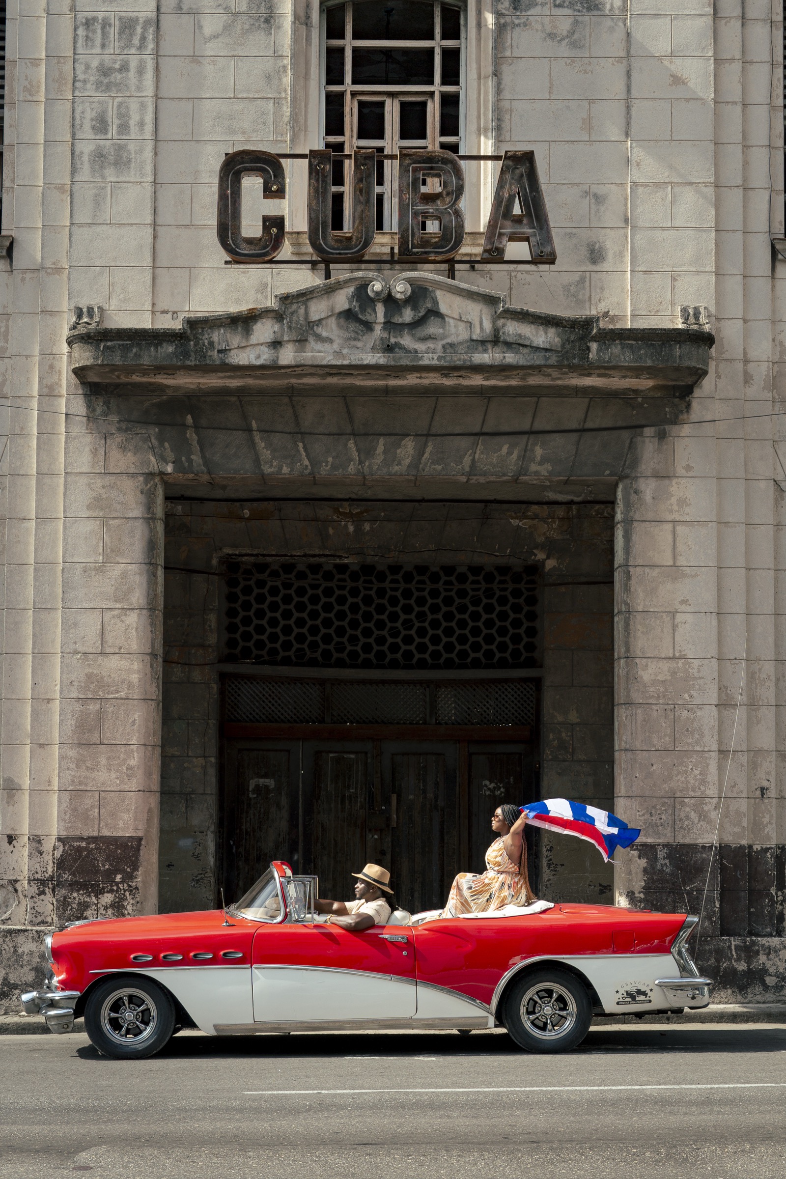 A couple in a classic car waving a Cuban flag in front of the cinema