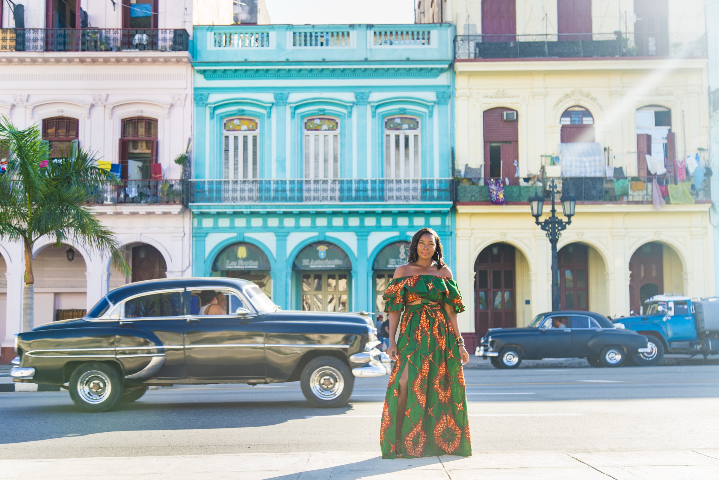 A woman in a patterned dress on the road, a classic car passing behind pastel facades