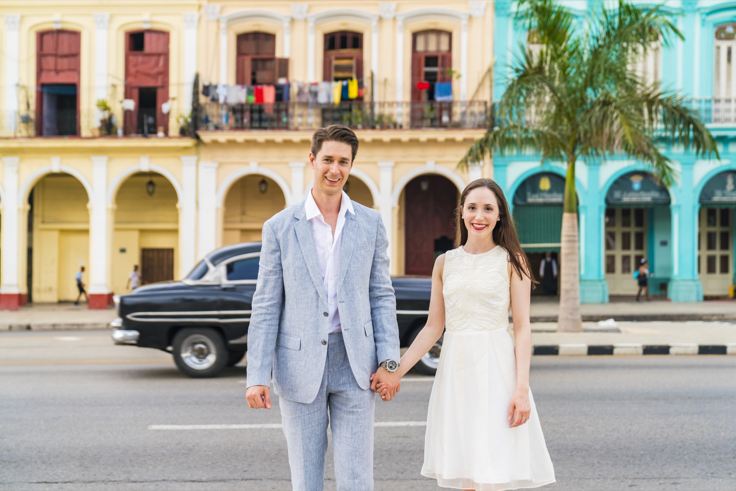 A couple holding hands, a black classic car crossing behind the pastel arcades