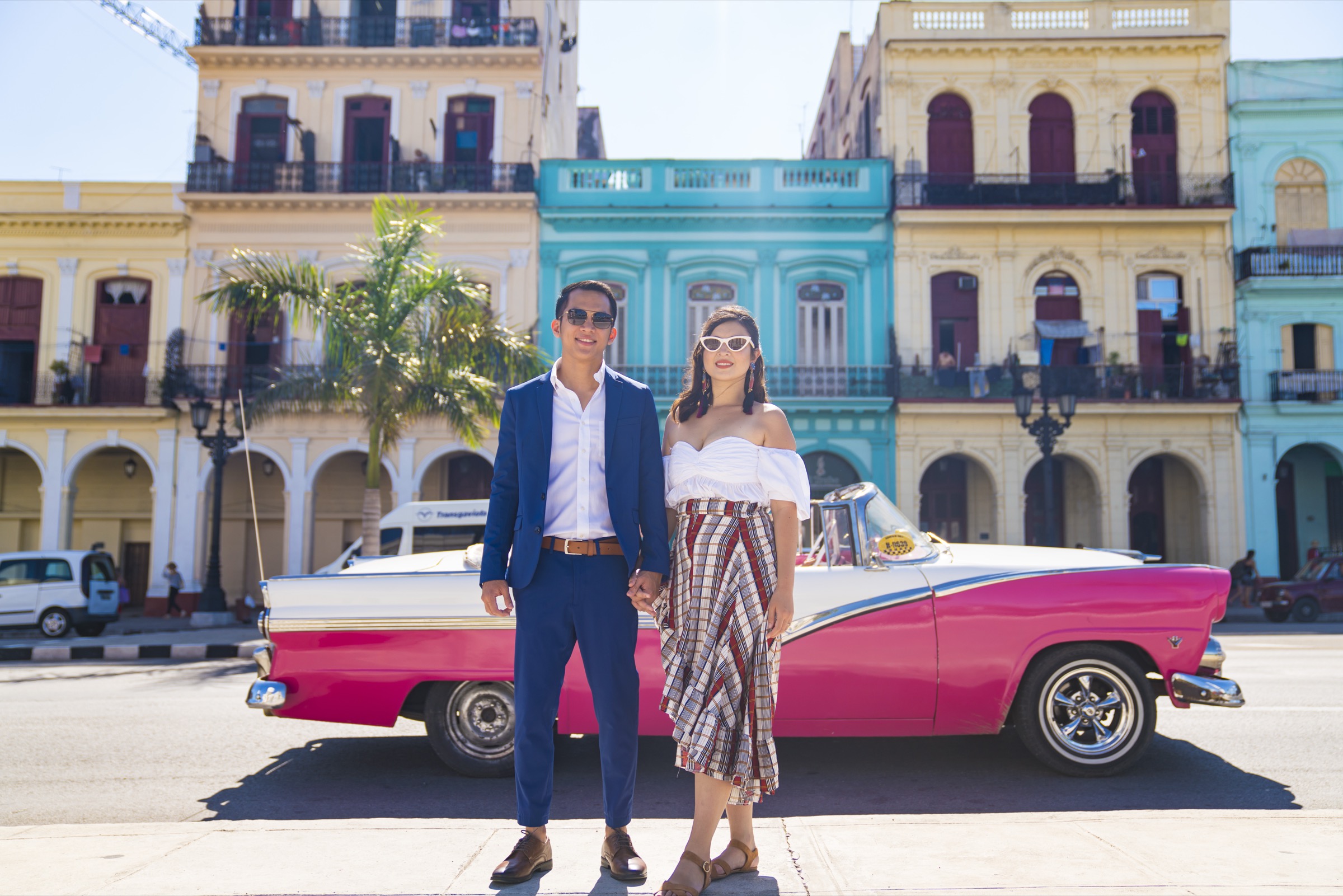 A couple posed beside a pink classic convertible, pastel houses behind