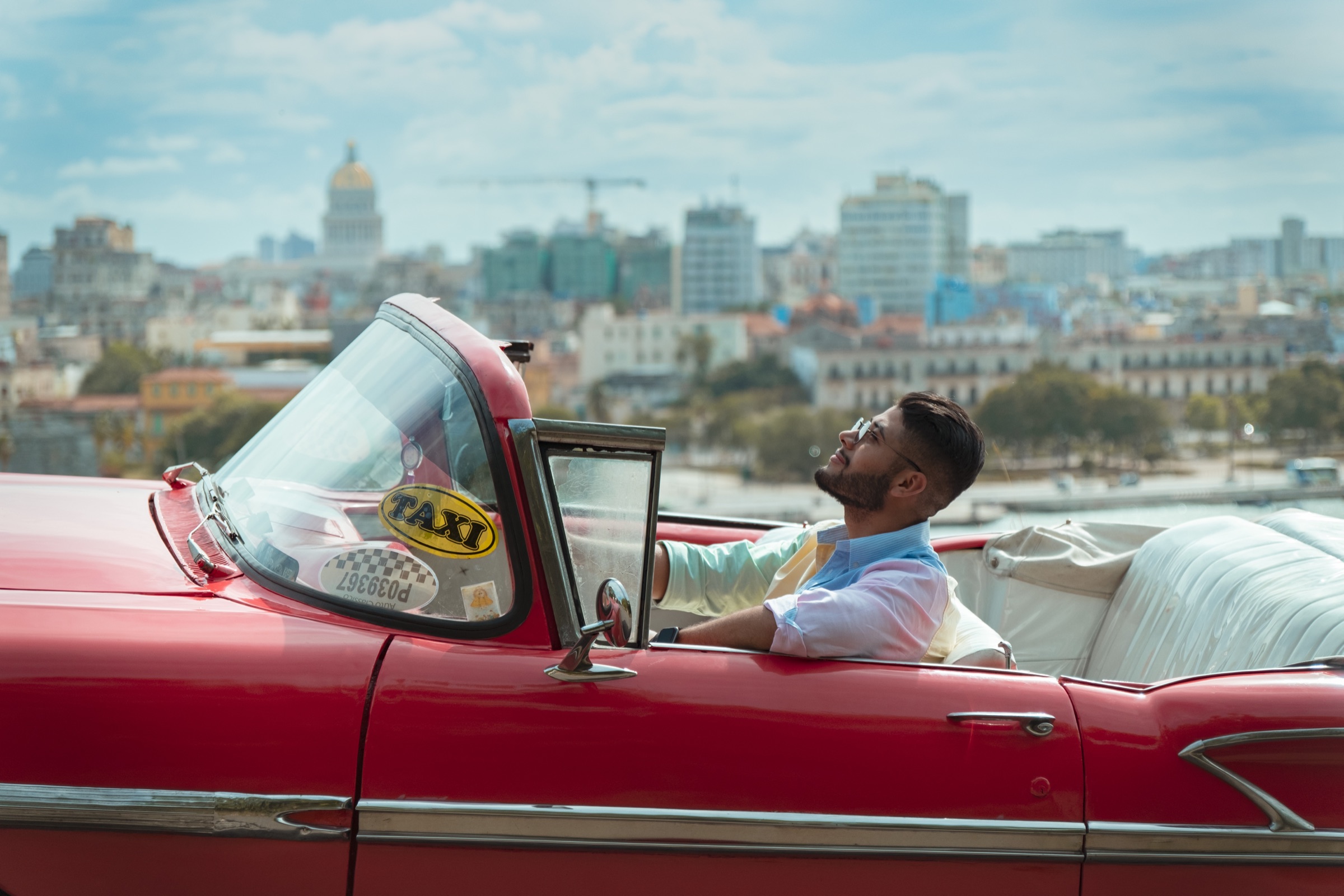 A man in a red classic convertible with the Havana skyline blurred behind him