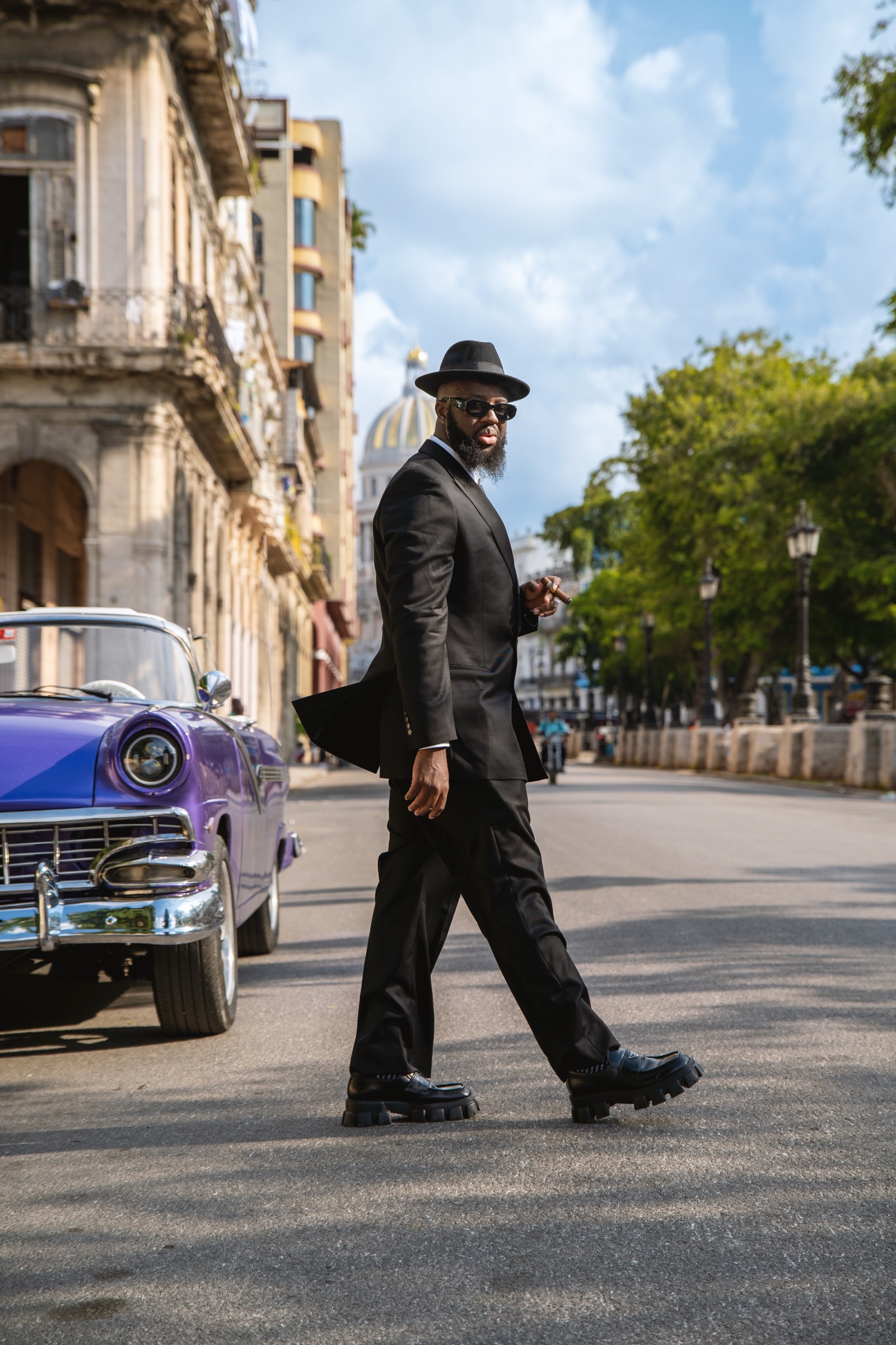A man in a black suit walking down the boulevard, a purple classic car beside him
