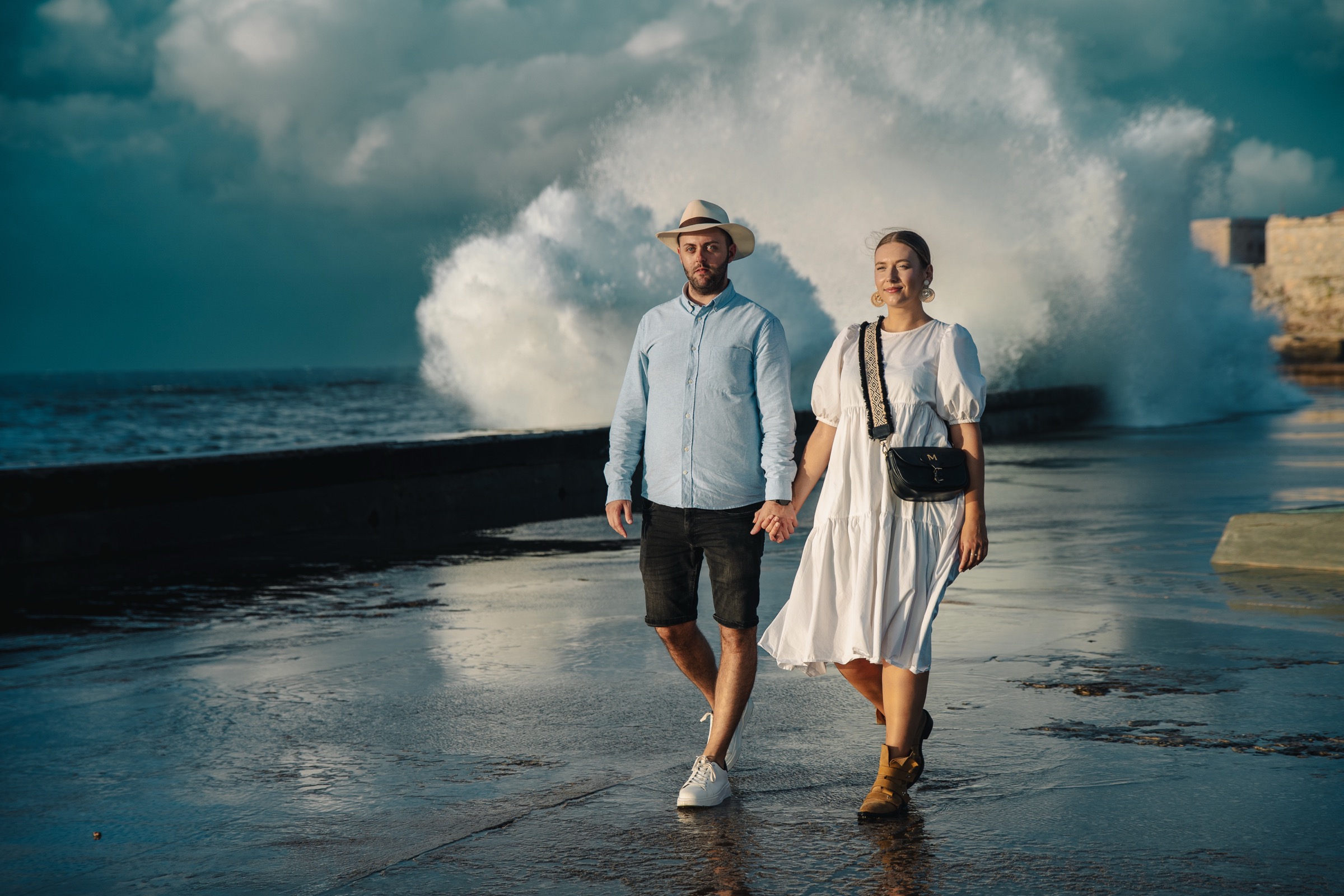 A couple walking the wet seawall road, a huge wave crashing behind them