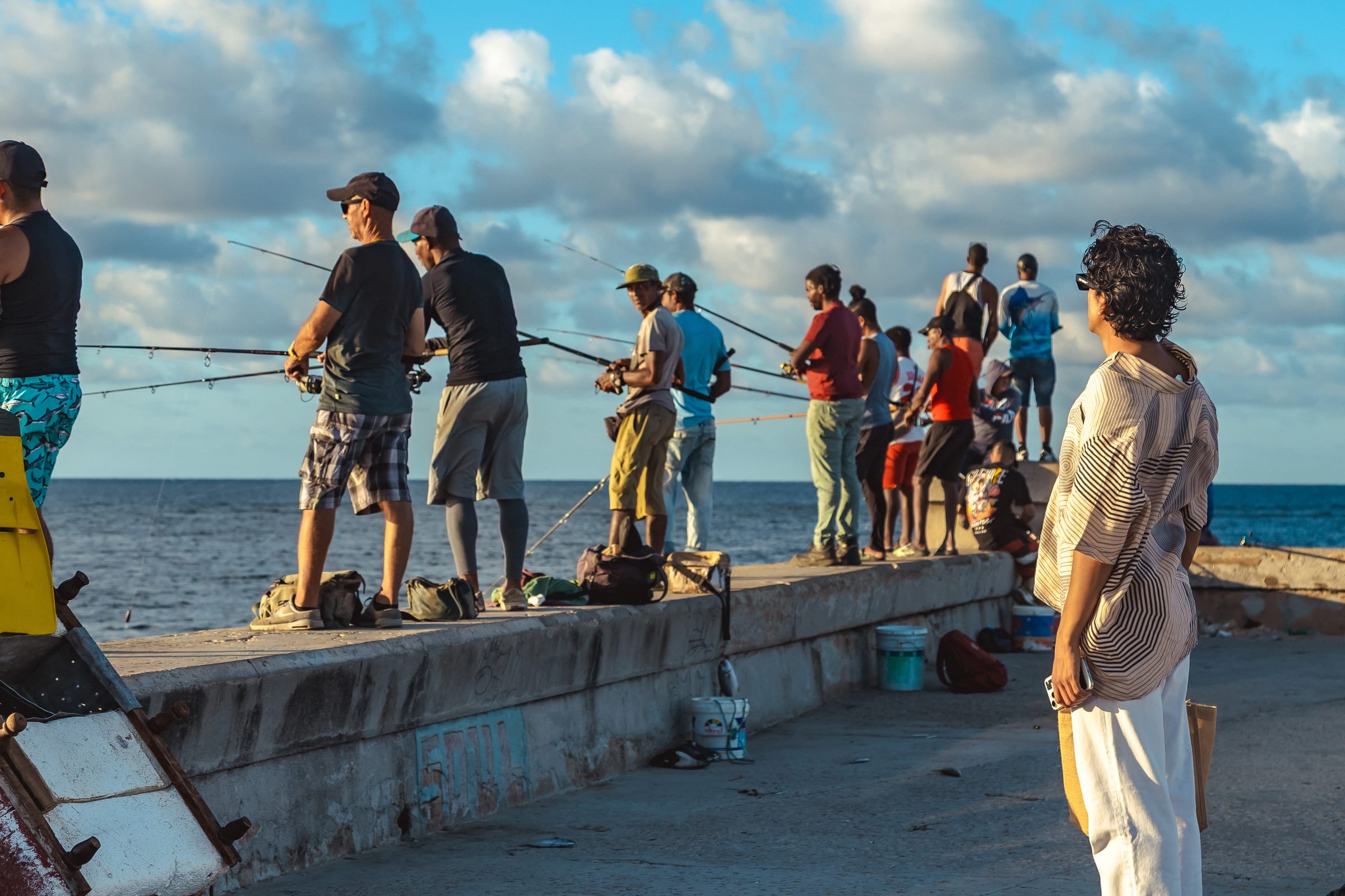 A row of fishermen casting from the Malecón seawall at golden hour, a woman watching them