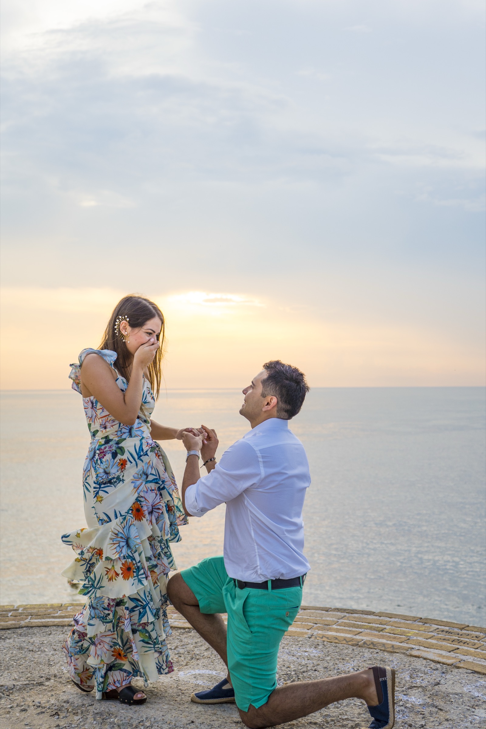 A man kneeling to propose, the woman with hands to her face, sunset over the sea