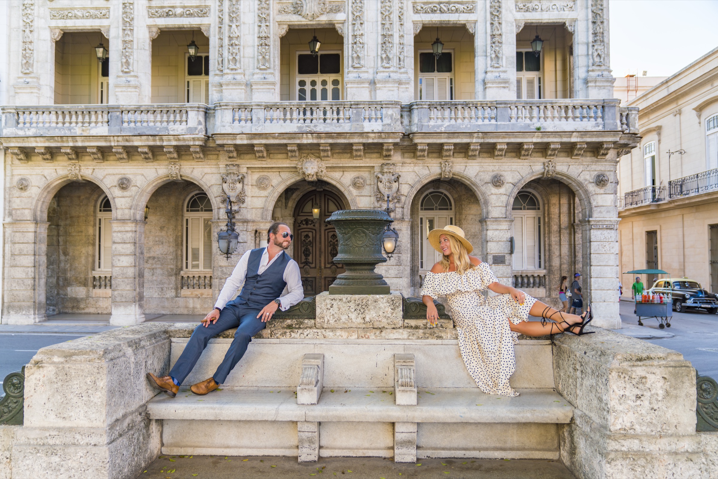 A couple seated either side of a bronze urn on a marble bench