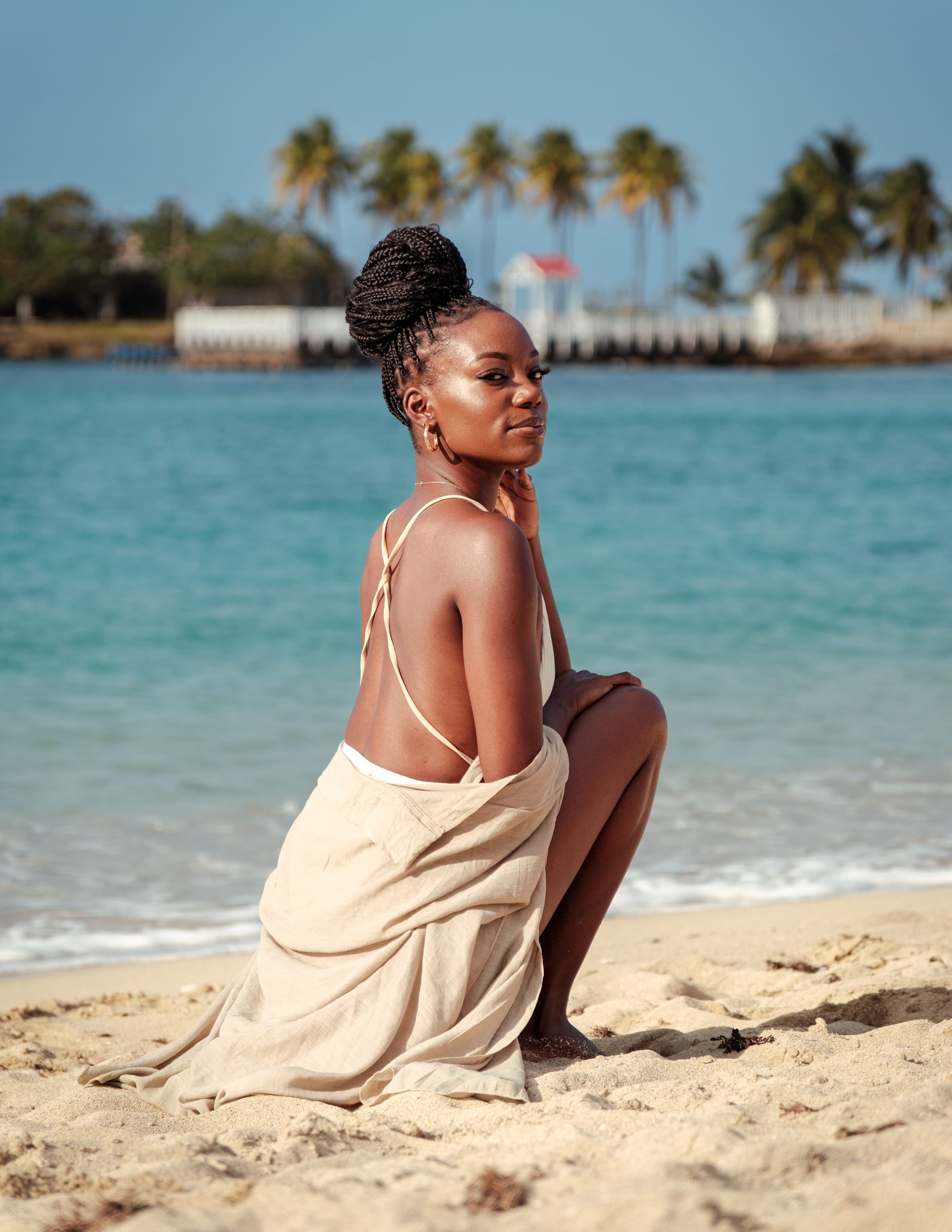 A woman in a beige linen dress kneeling on the white sand with palms and the old pier soft behind her