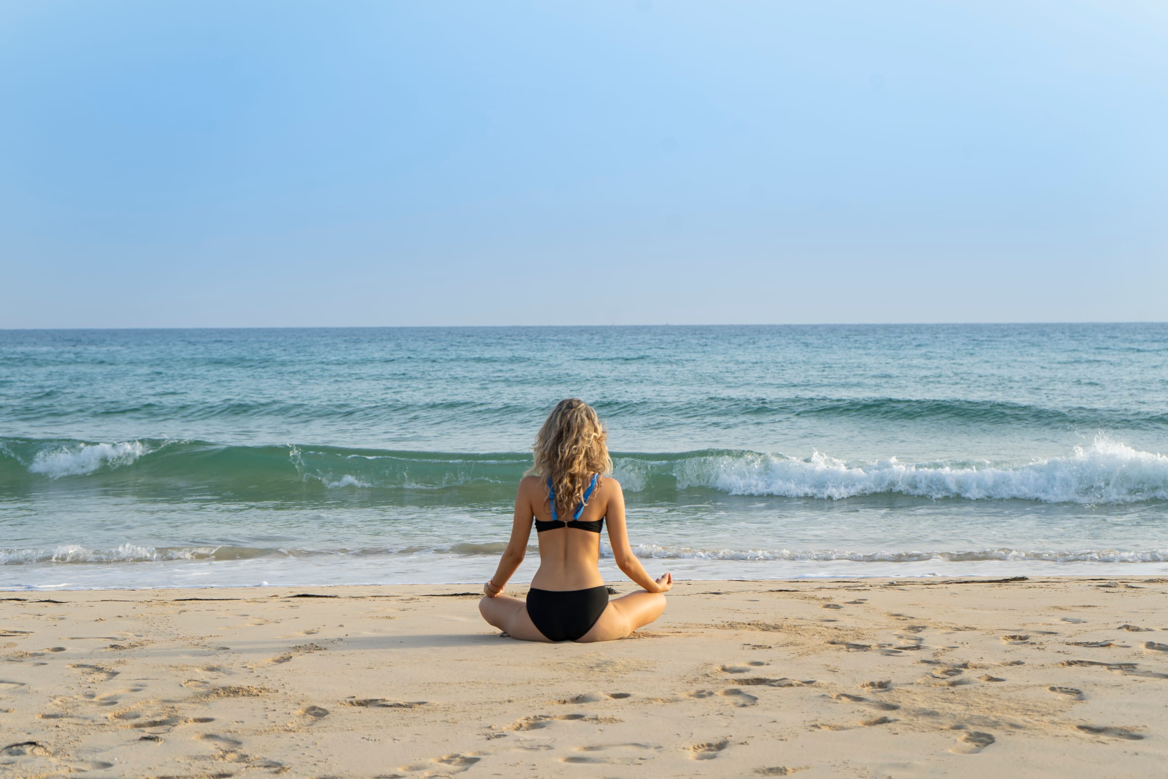 A woman seated cross-legged at the shoreline facing the waves in early light