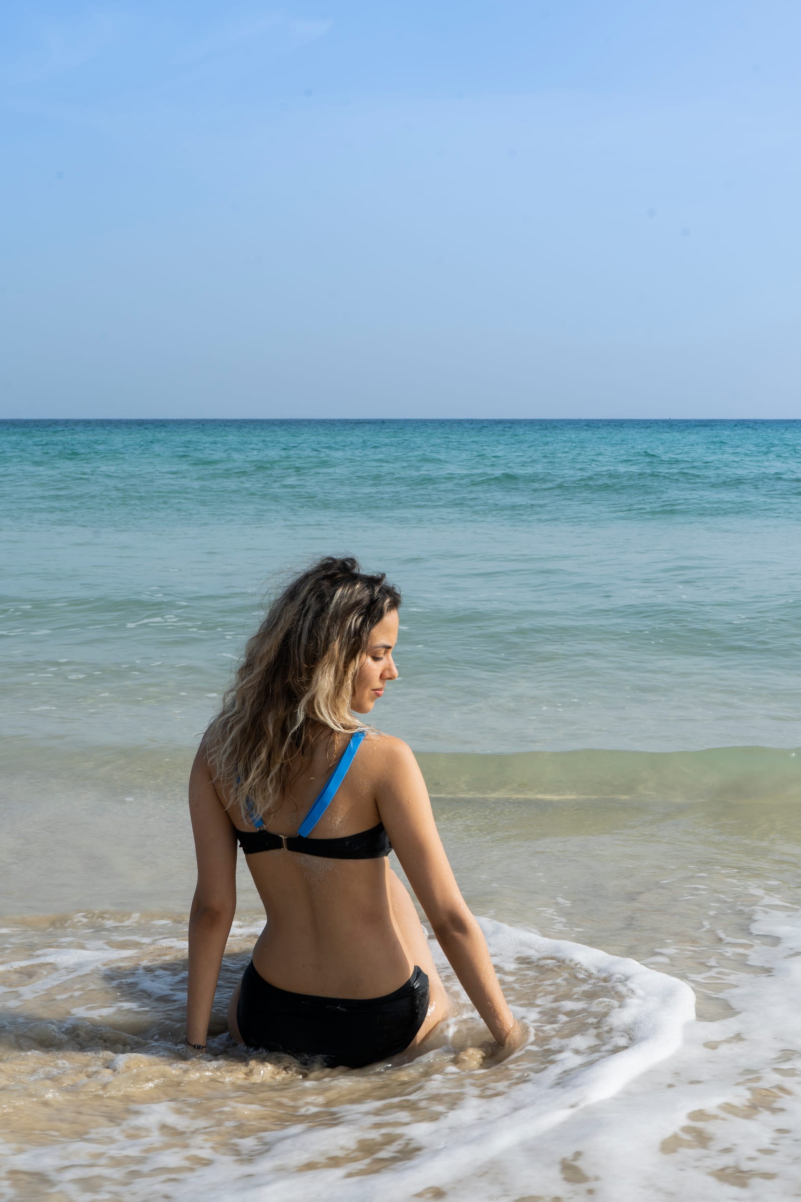 A woman in a black swimsuit sitting in the shallow wash, looking down along the shore