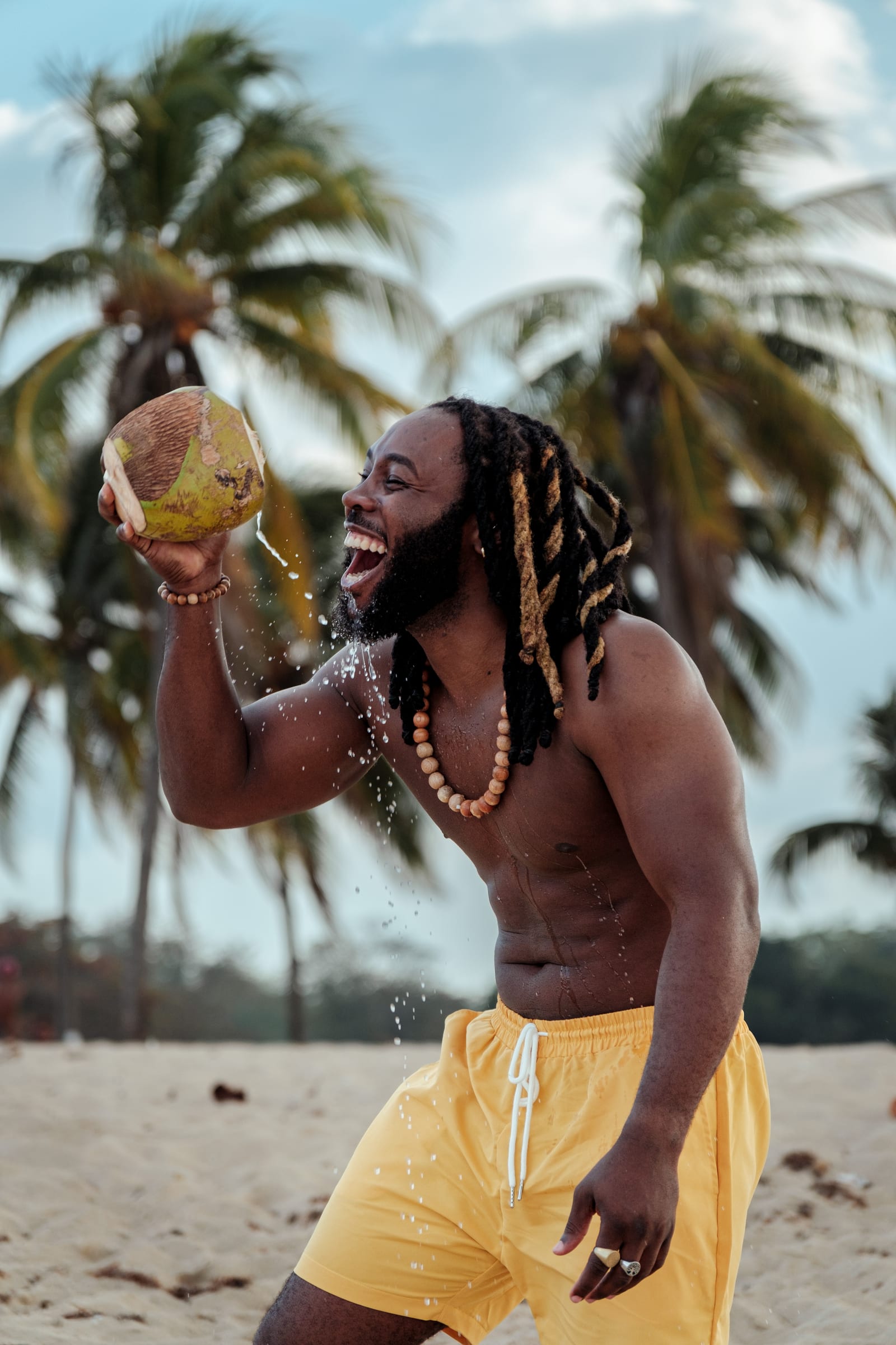 A man laughing as water splashes from a fresh green coconut, palm trees behind him